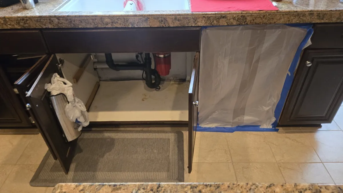 Under-sink cabinet water damage before remediation showing stained cabinet floor and containment plastic